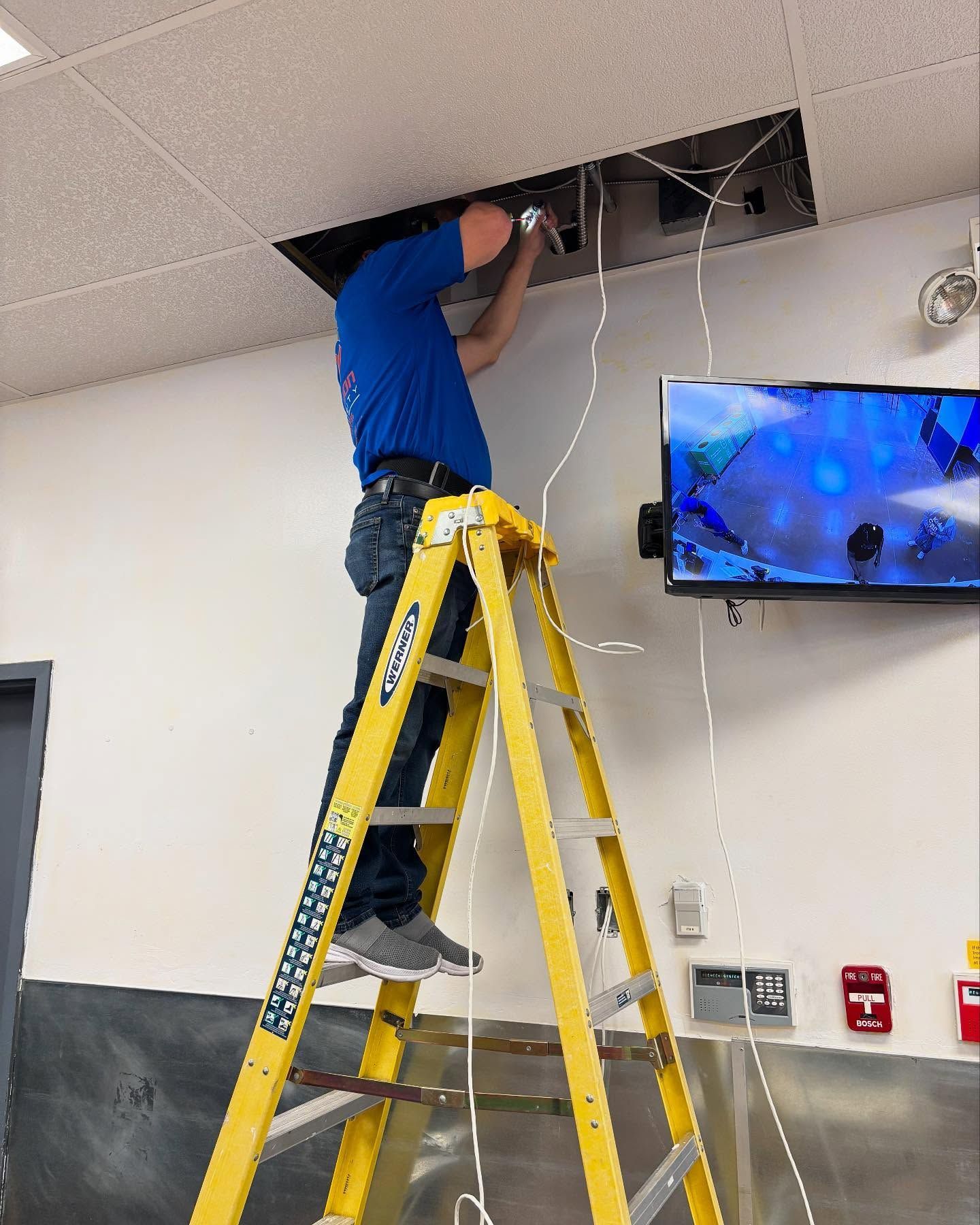 Man on a yellow ladder installing wires into a ceiling panel, near a wall-mounted TV.