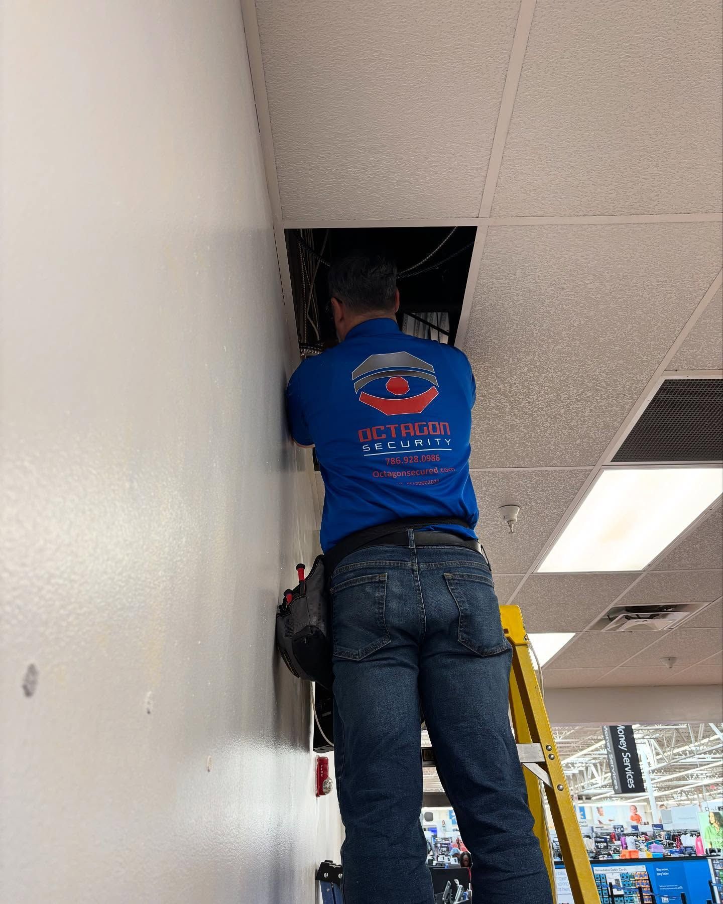 Man on a ladder reaching into a ceiling panel, likely doing electrical work. Blue shirt, tool belt.
