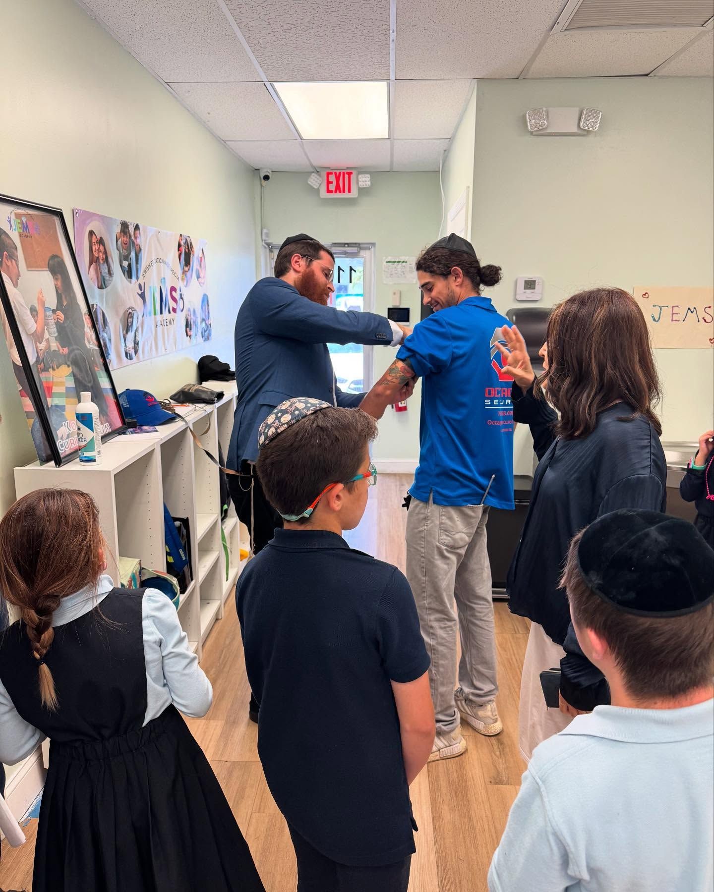 A man in a suit measuring a person's arm, surrounded by people, in a hallway.