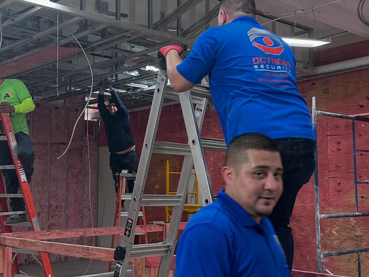 Construction workers installing wiring in a building. Two on ladders, one on the ground.