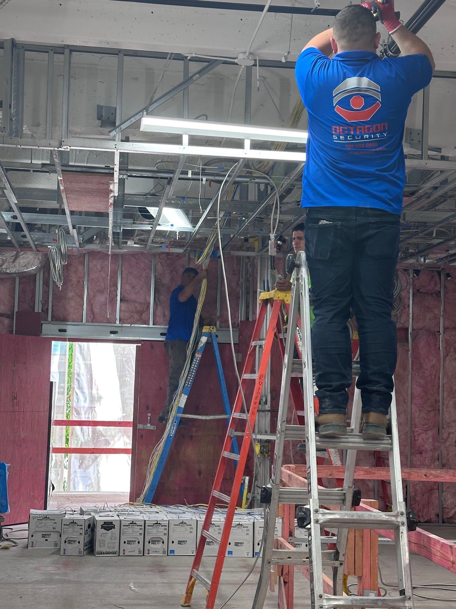 Construction workers installing ceiling fixtures, standing on ladders. Interior setting with exposed framework.
