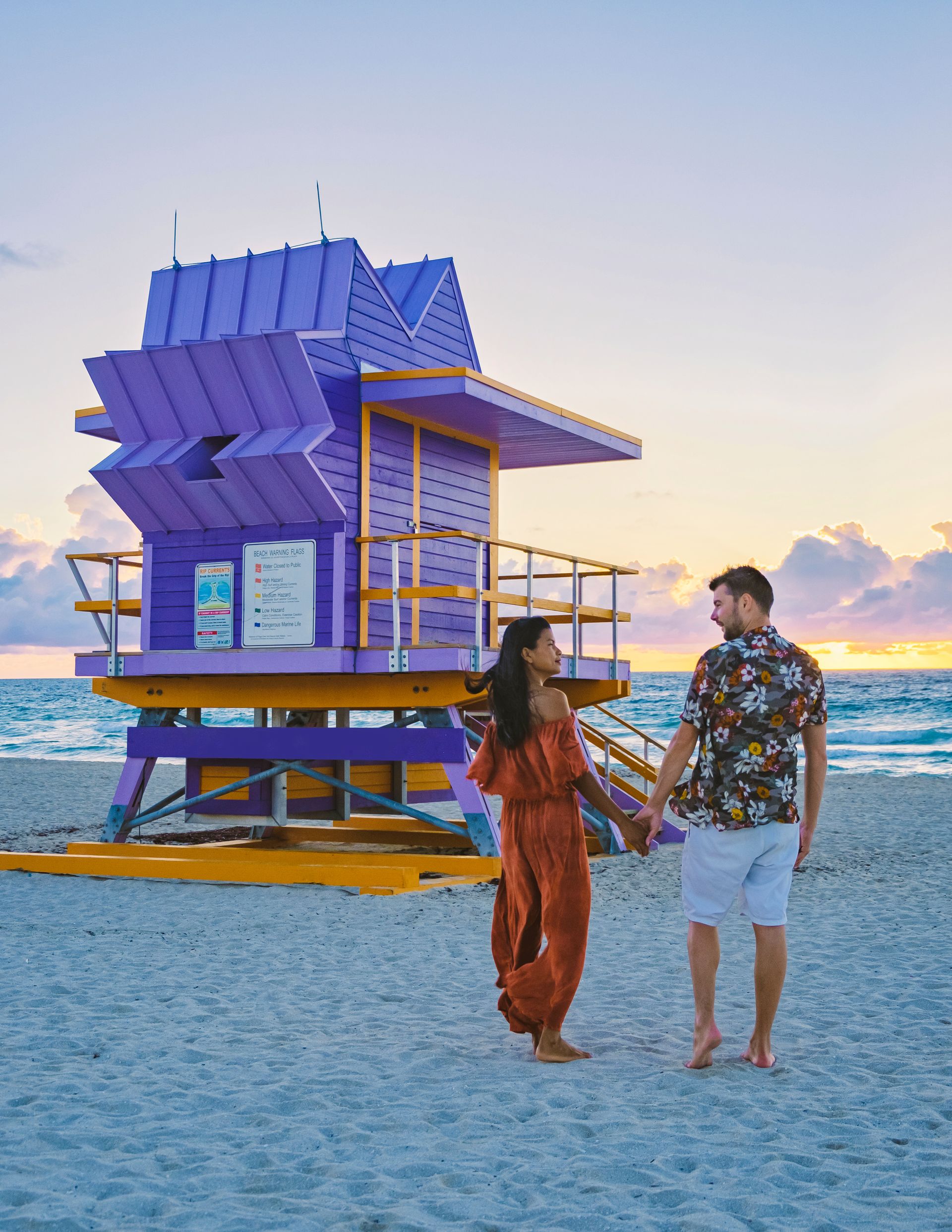 A man and a woman are holding hands on the beach in front of a purple lifeguard tower.