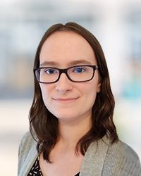 Woman wearing glasses smiles at the camera, light skin, brown hair, wearing a gray sweater.