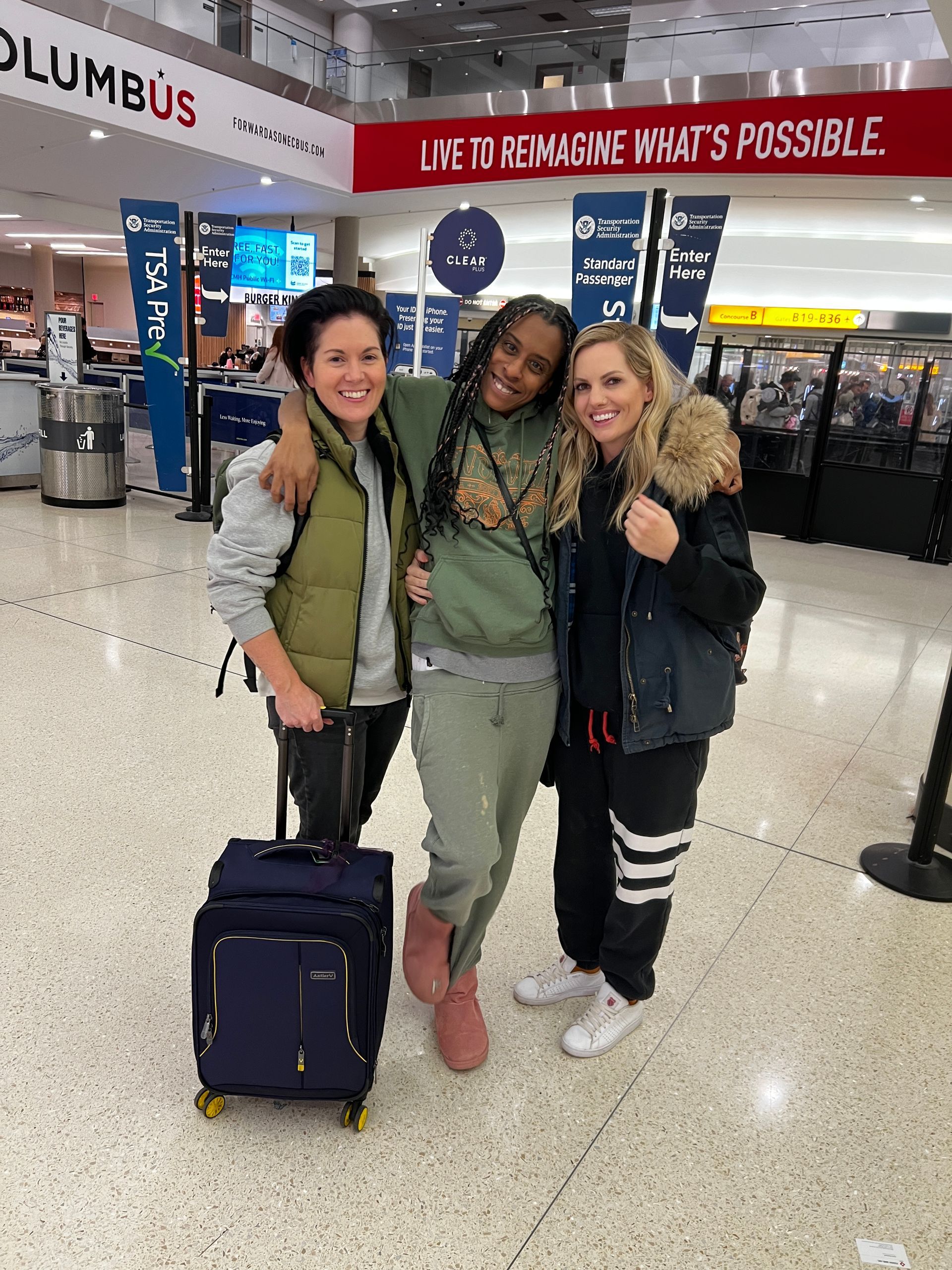 Three women are posing for a picture in an airport with luggage.