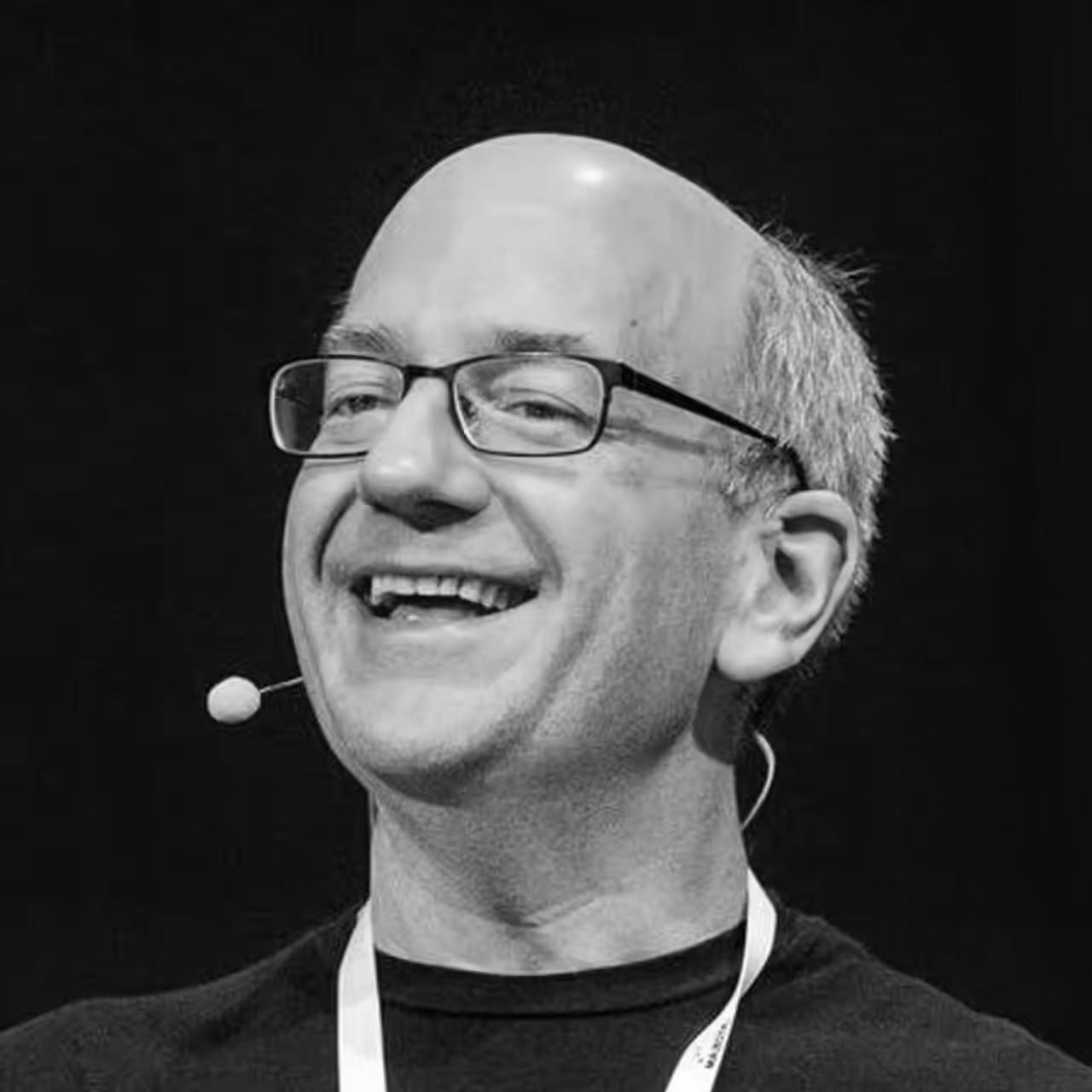 A man wearing glasses and a microphone is smiling in a black and white photo.