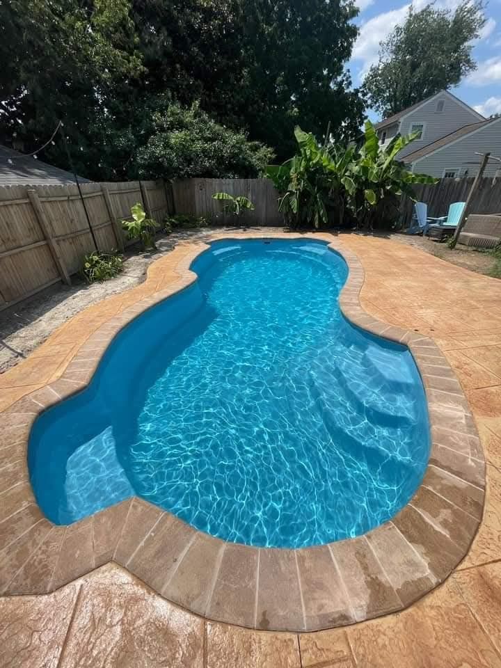 A backyard swimming pool with blue water and steps, surrounded by stamped concrete and a fence.