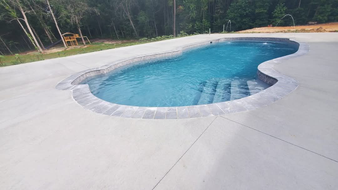 Swimming pool with concrete deck and steps leading into the blue water. Forest in background.