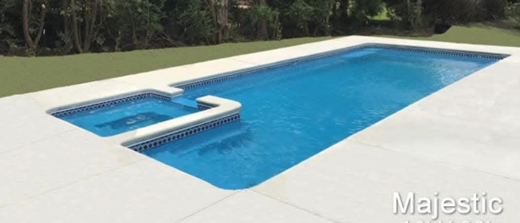 Swimming pool with a connected spa surrounded by white tiles and green grass.
