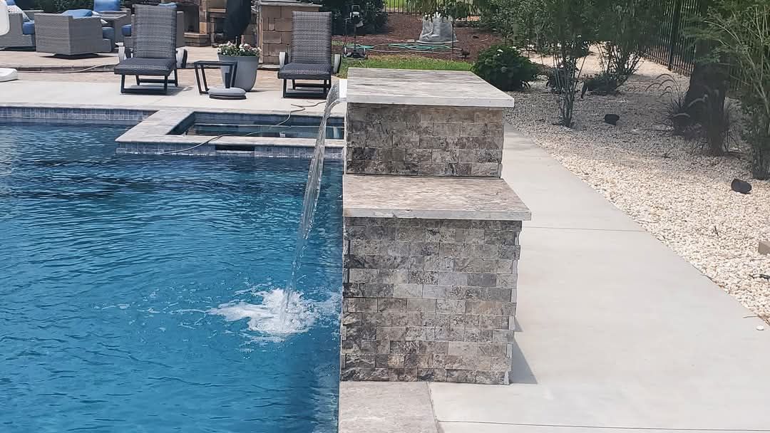 Poolside waterfall feature; stone wall with water cascading into pool; lounge chairs in background.
