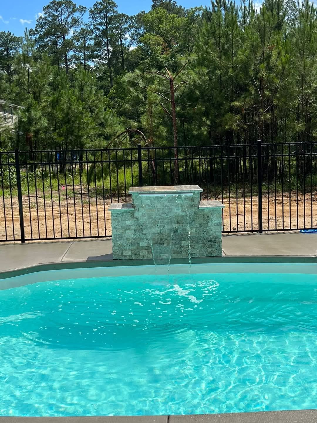 Pool with waterfall feature and black fence, surrounded by trees under a blue sky.
