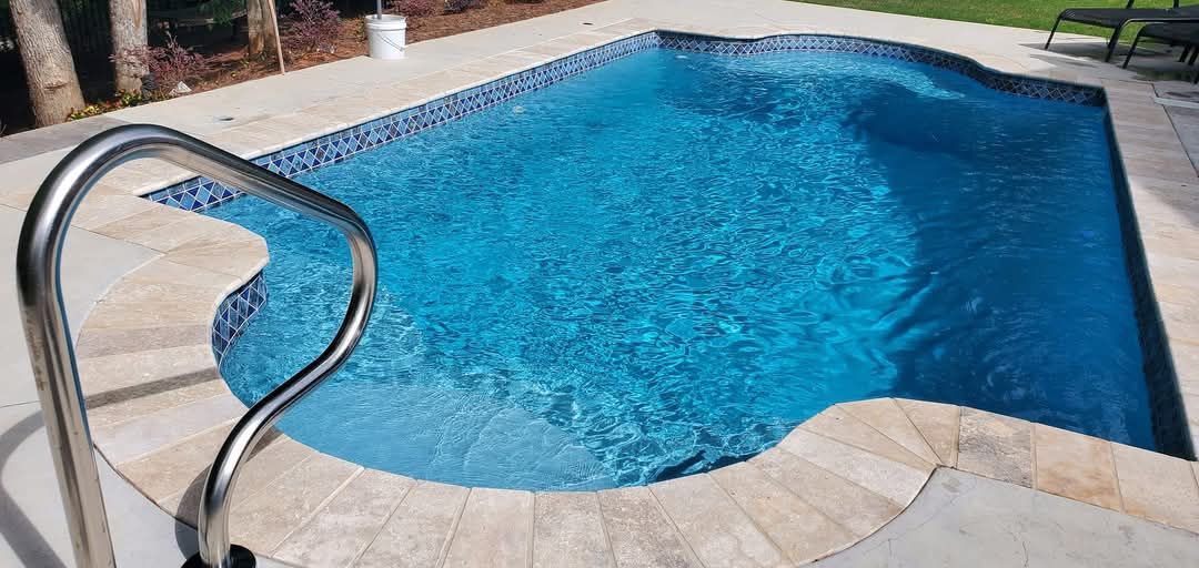 A clear blue swimming pool with a curved edge and a silver ladder. Light beige tiles surround the pool.