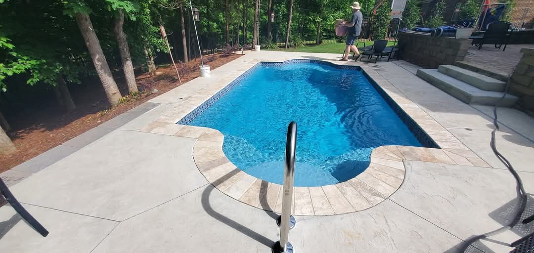 A blue pool with a handrail surrounded by concrete. Trees and a person are in the background.
