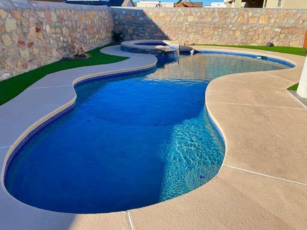Swimming pool with a curved shape, surrounded by light-colored concrete and stone wall.