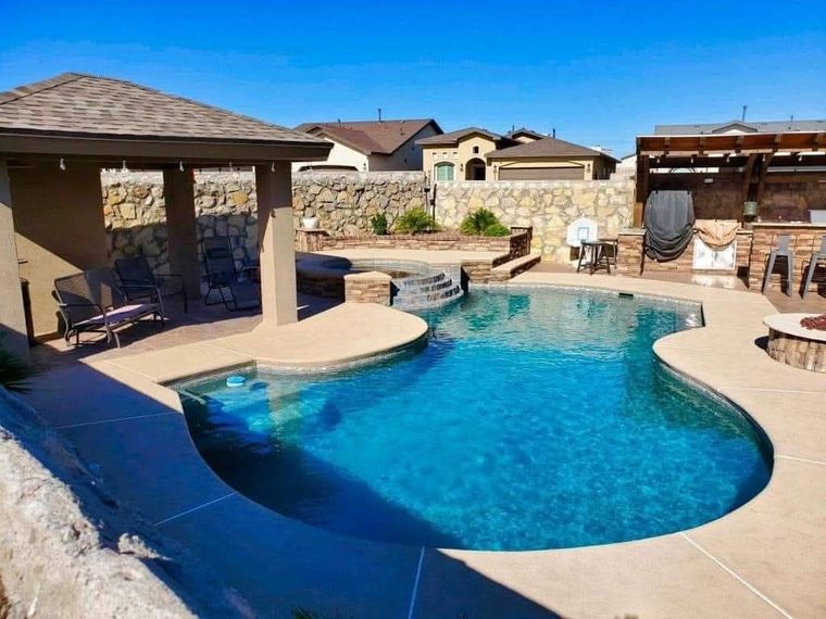 Swimming pool in a backyard with a gazebo, outdoor kitchen, and stone wall under a clear sky.