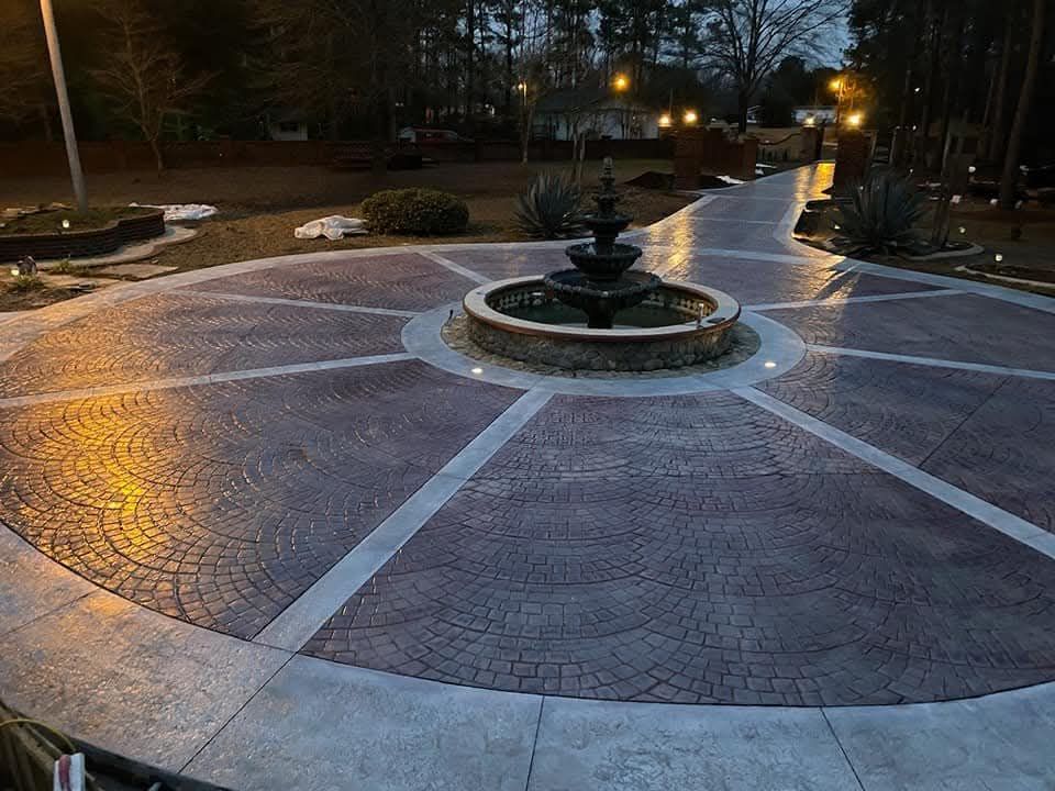 Concrete driveway with fountain at dusk, illuminated by lights.