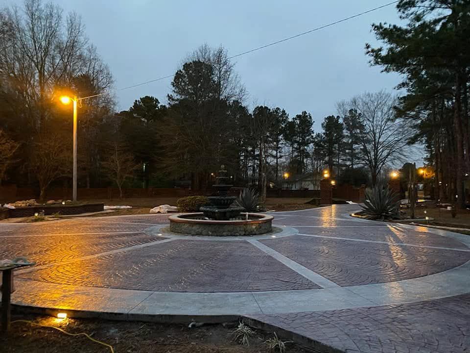 Fountain in a brick plaza under overcast sky, surrounded by trees and decorative lighting.