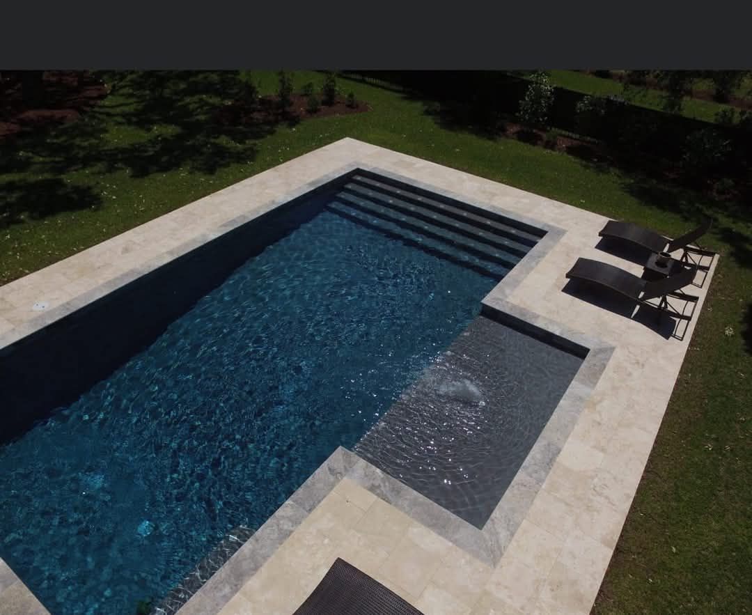 Overhead view of a rectangular pool with dark blue water and stone patio surrounded by green grass.