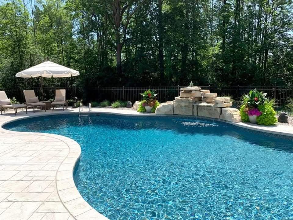 Swimming pool with waterfall, chairs, and umbrella in a backyard. Blue water, stone patio, green trees.