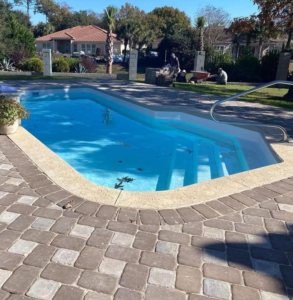 Swimming pool with steps, surrounded by stone patio, green lawn, and a house.