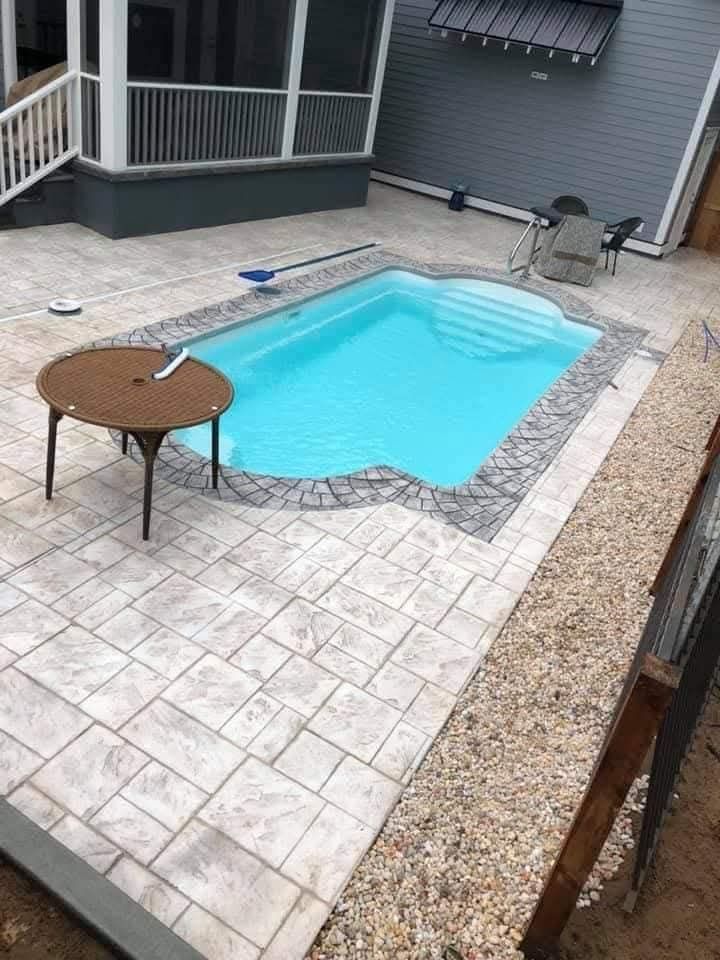 Small backyard pool with stone patio, round table, and pebble border.
