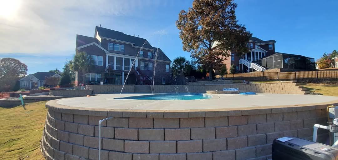 A backyard with a pool, brick retaining walls, and large houses under a blue sky.