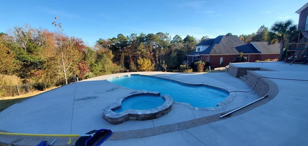 A large outdoor pool and spa surrounded by a concrete patio, with a house and trees in the background.