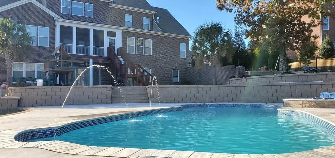 A backyard swimming pool with fountains, a multi-story brick house, and tiered retaining walls.
