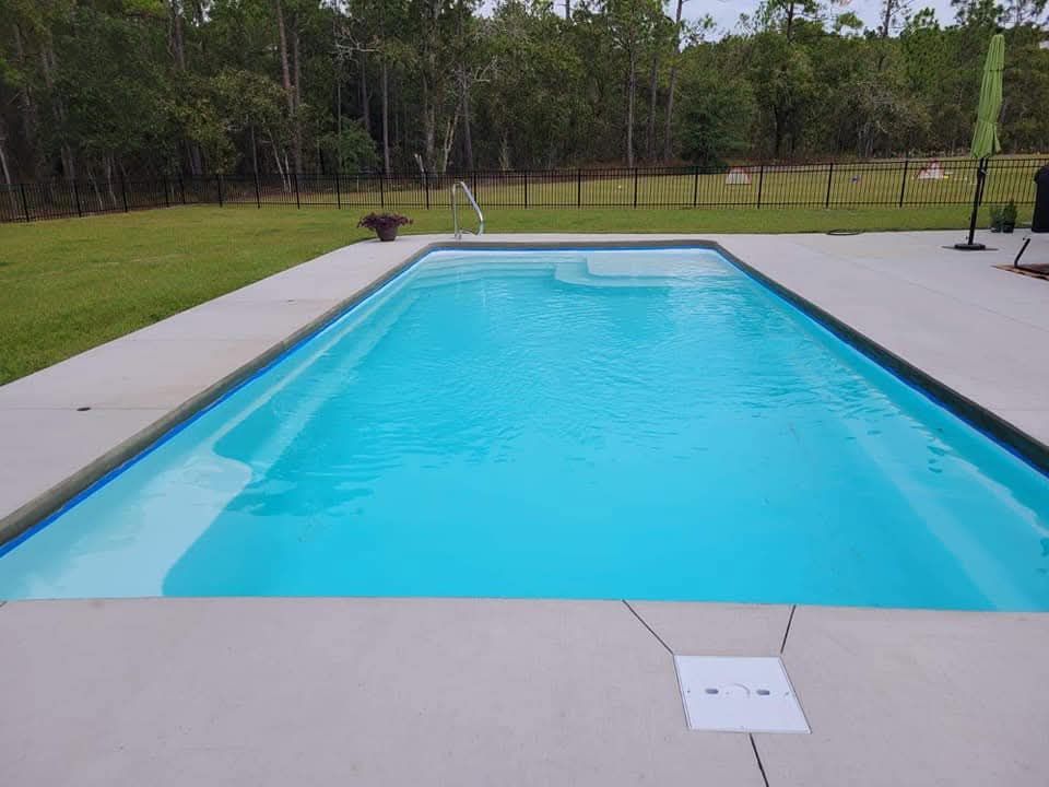 Rectangular swimming pool with bright blue water surrounded by concrete patio. Green lawn and trees in the background.