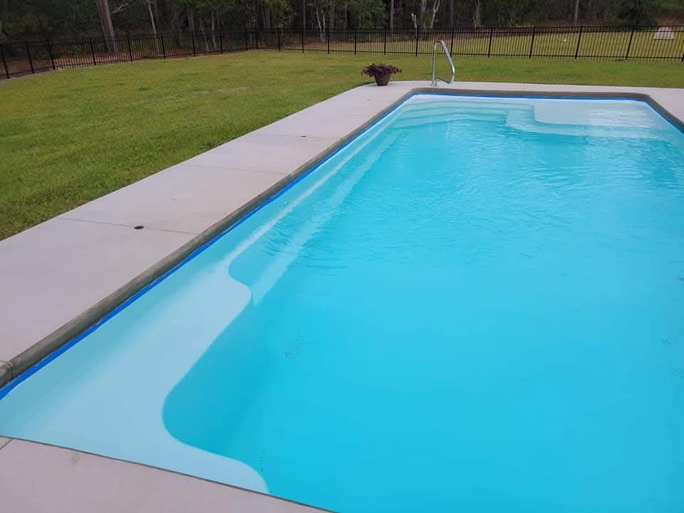 Swimming pool with light blue water and a concrete border, set in a grassy backyard.