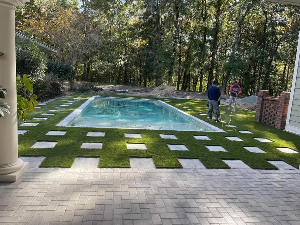 Pool area with pavers, artificial turf squares, and two people working in a wooded setting.