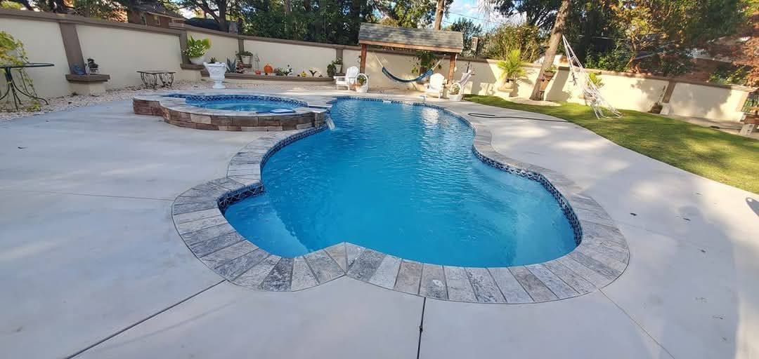 A blue swimming pool with a stone border in a backyard. A hot tub is nearby.