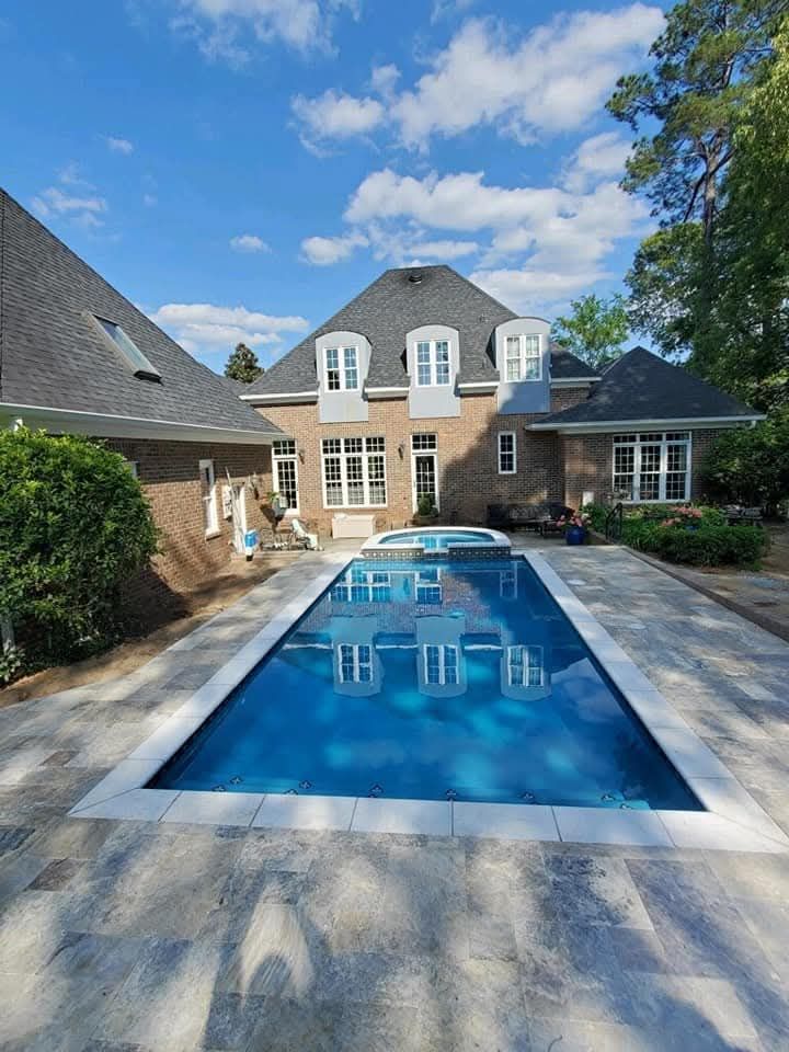 Backyard with rectangular pool, light gray stone patio, and brick house under blue sky.