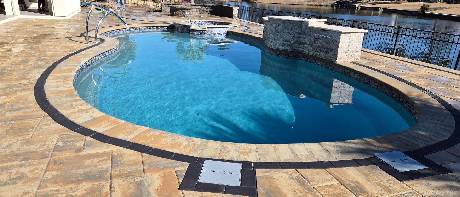 Oval-shaped pool with blue water. Stone border with patio, near a lake.
