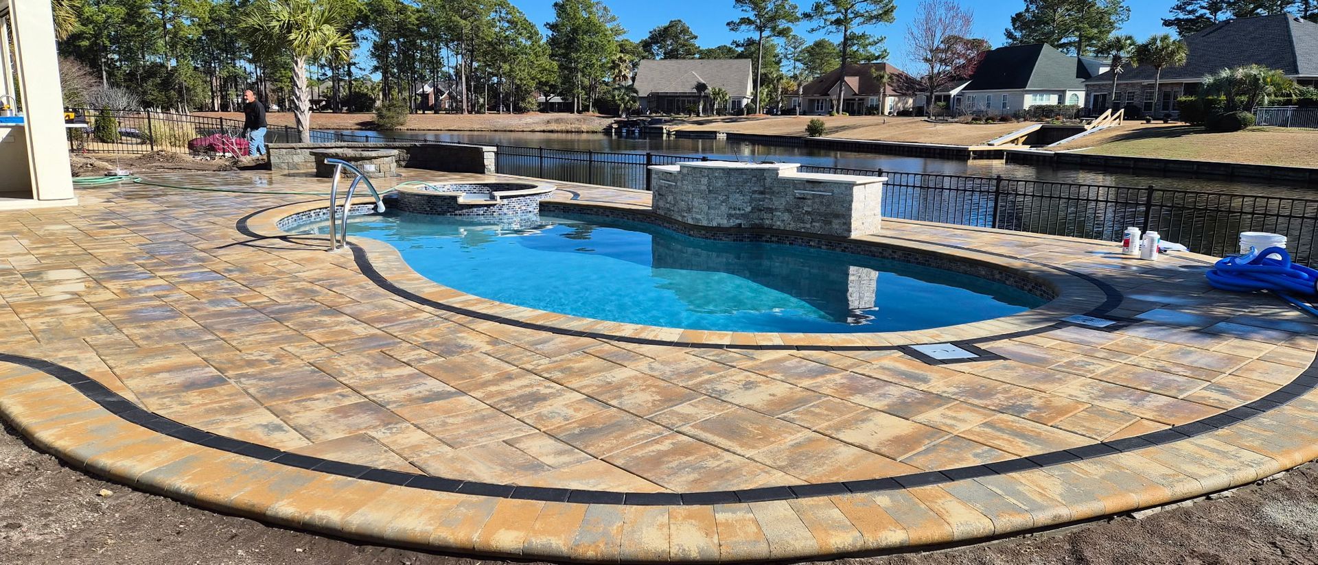 A pool with turquoise water surrounded by brick-like patio, a lake is in the background.