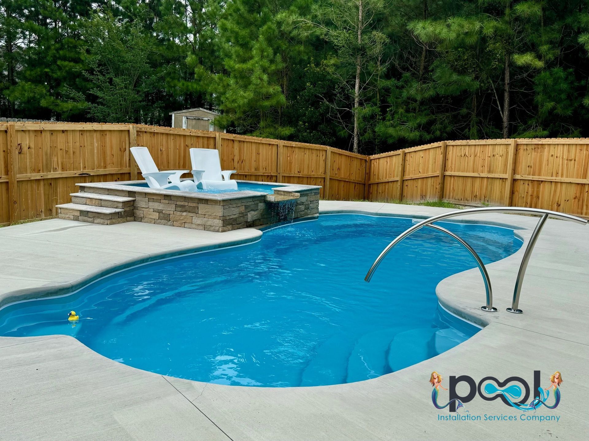 Swimming pool with spa, concrete deck, wooden fence, white chairs, blue water.