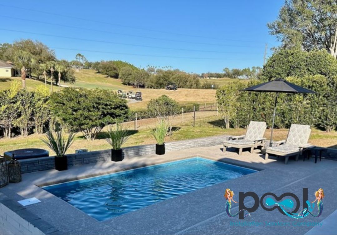 A rectangular pool with lounge chairs and a black umbrella overlooking a grassy landscape under a blue sky.