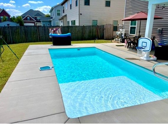 Rectangular blue swimming pool in a backyard, concrete patio, jacuzzi, and shade umbrella visible.