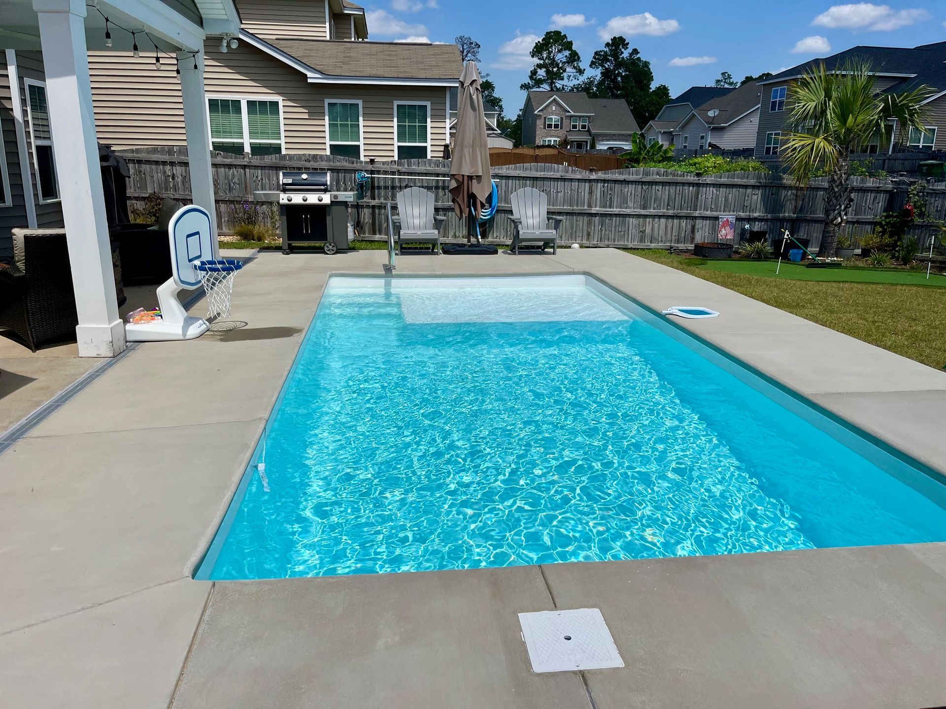 Rectangular backyard pool with blue water. Concrete patio, grill, basketball hoop, and chairs.