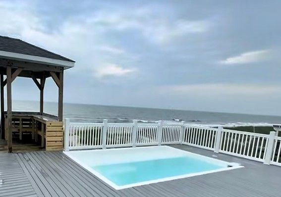 Wooden deck with pool, gazebo, and white railing overlooking the ocean under a cloudy sky.