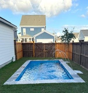 Small rectangular backyard pool with a water fountain, surrounded by grass and a wooden fence.