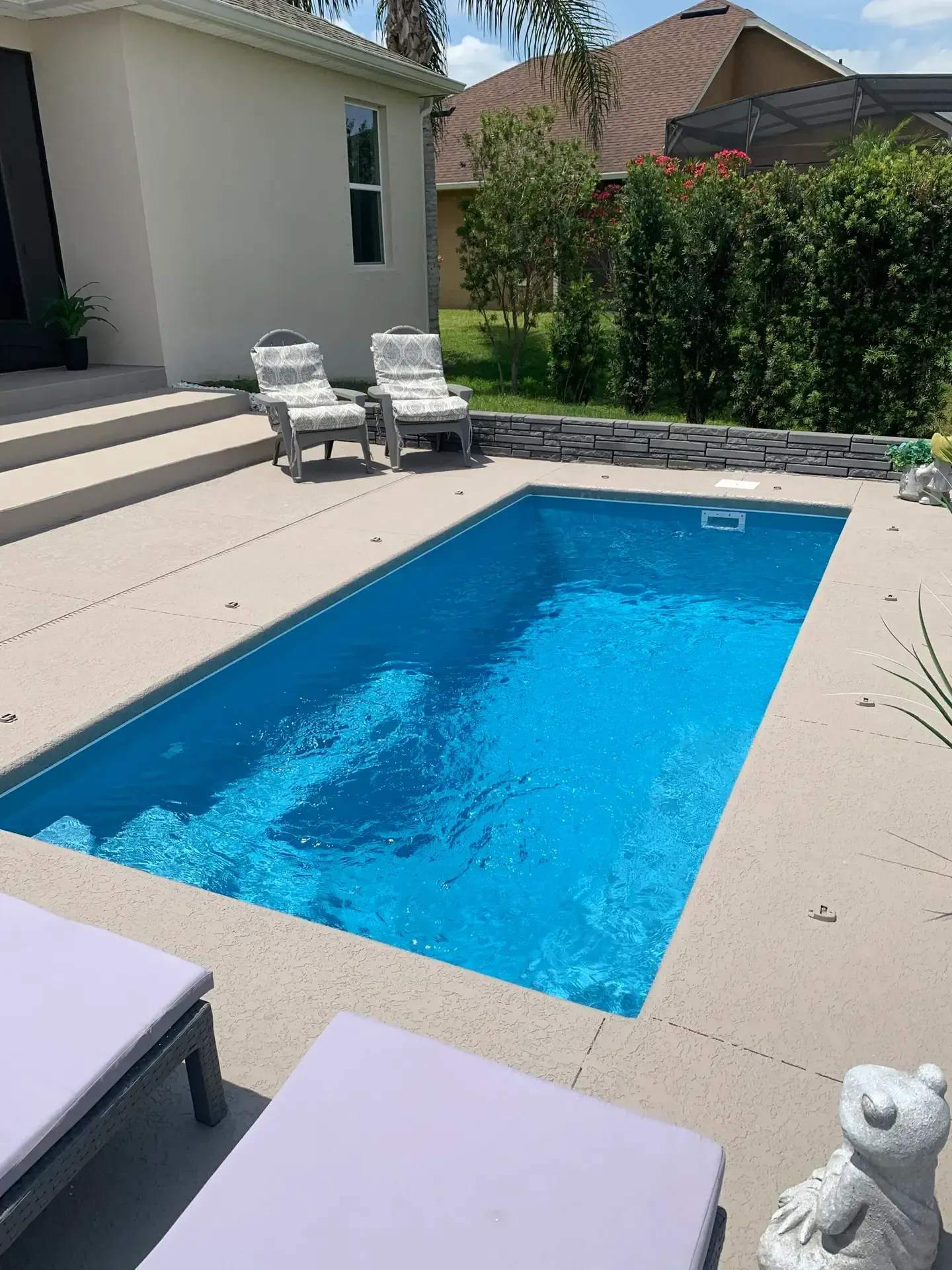 Small rectangular pool with blue water surrounded by concrete patio, lounge chairs, and landscaping.
