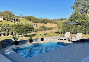 Pool with lounge chairs on a patio, overlooking a green field under a clear blue sky.