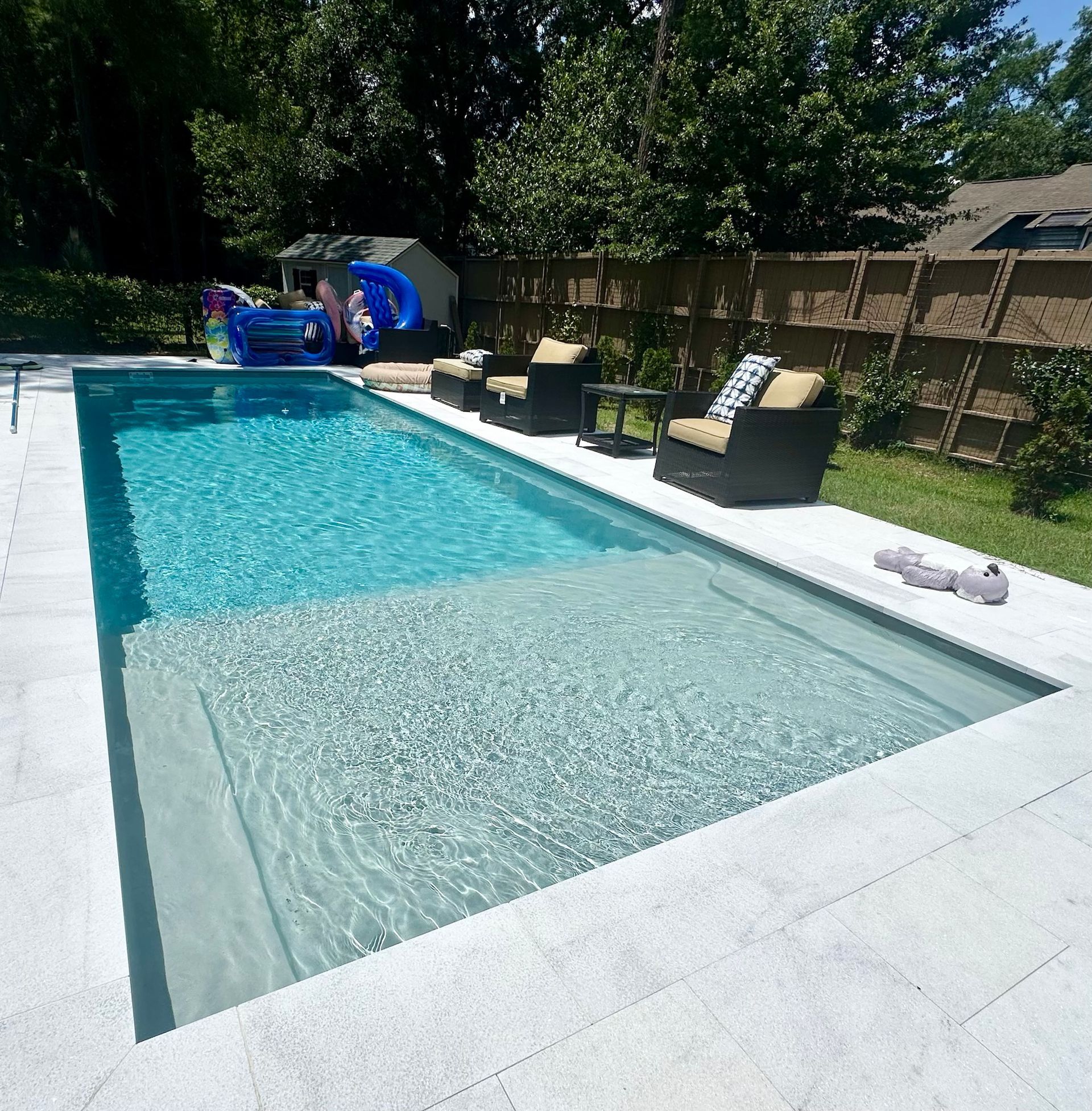 Rectangular pool with shallow and deep ends; chairs and a fence in the background. Blue water and light gray surroundings.