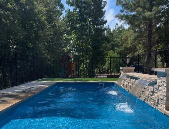 Pool with blue water and waterfall feature, surrounded by greenery and a black fence.