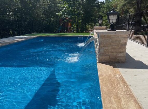 Blue swimming pool with water feature, stone wall, and black lamp posts. Sunny outdoor setting.