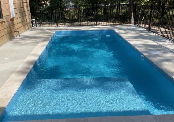 Rectangular swimming pool with blue water surrounded by light-colored concrete and a black fence.