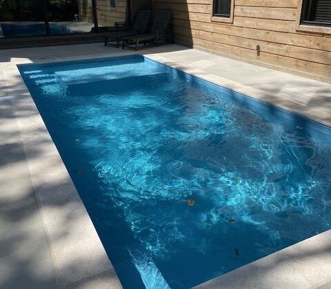 Rectangular outdoor pool with light blue water and surrounding light-colored stone patio, near a wooden building.