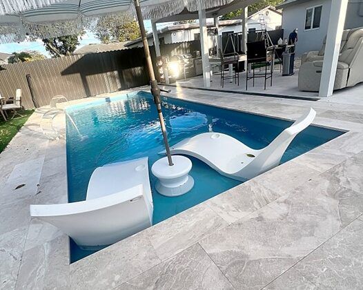 A backyard pool with lounge chairs and an umbrella, surrounded by light gray stone.