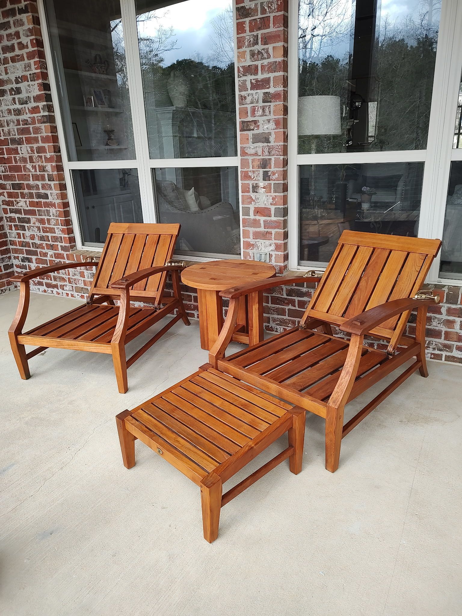 Two wooden lounge chairs and a small table on a porch with brick walls.