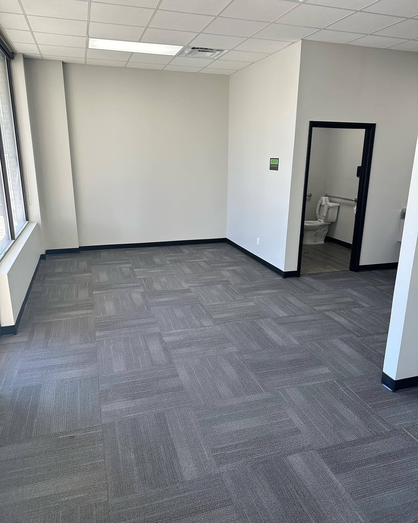 Empty office room with gray carpet, white walls, and a door to a bathroom.